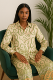 : Femme assise sur un fauteuil vert portant un ensemble en satin beige à motifs verts et orangés, chemise longue et pantalon fluide.