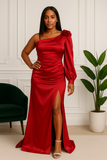 Femme debout en robe longue satinée rouge à une manche, avec fente haute et rosette sur l’épaule.